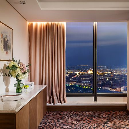 Grand Hotel Millennium 소피아 외부 사진 The photo depicts a modern hotel room or a lounge area with a stunning view of a cityscape at night. On the left, there's a wooden console table with a vase of flowers and a bottle of water. A piece of art is framed on the wall above the table. The r