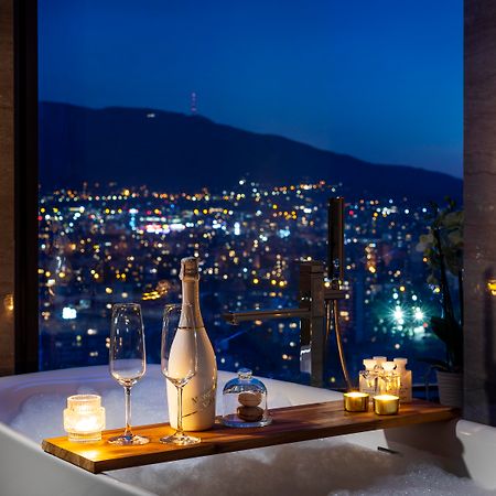 Grand Hotel Millennium 소피아 외부 사진 The photo features a luxurious bathtub placed by a large window, showcasing a scenic city view at night. There is a wooden tray resting on the edge of the tub, displaying a bottle of champagne, two glass flutes, and several lit candles, creating an a