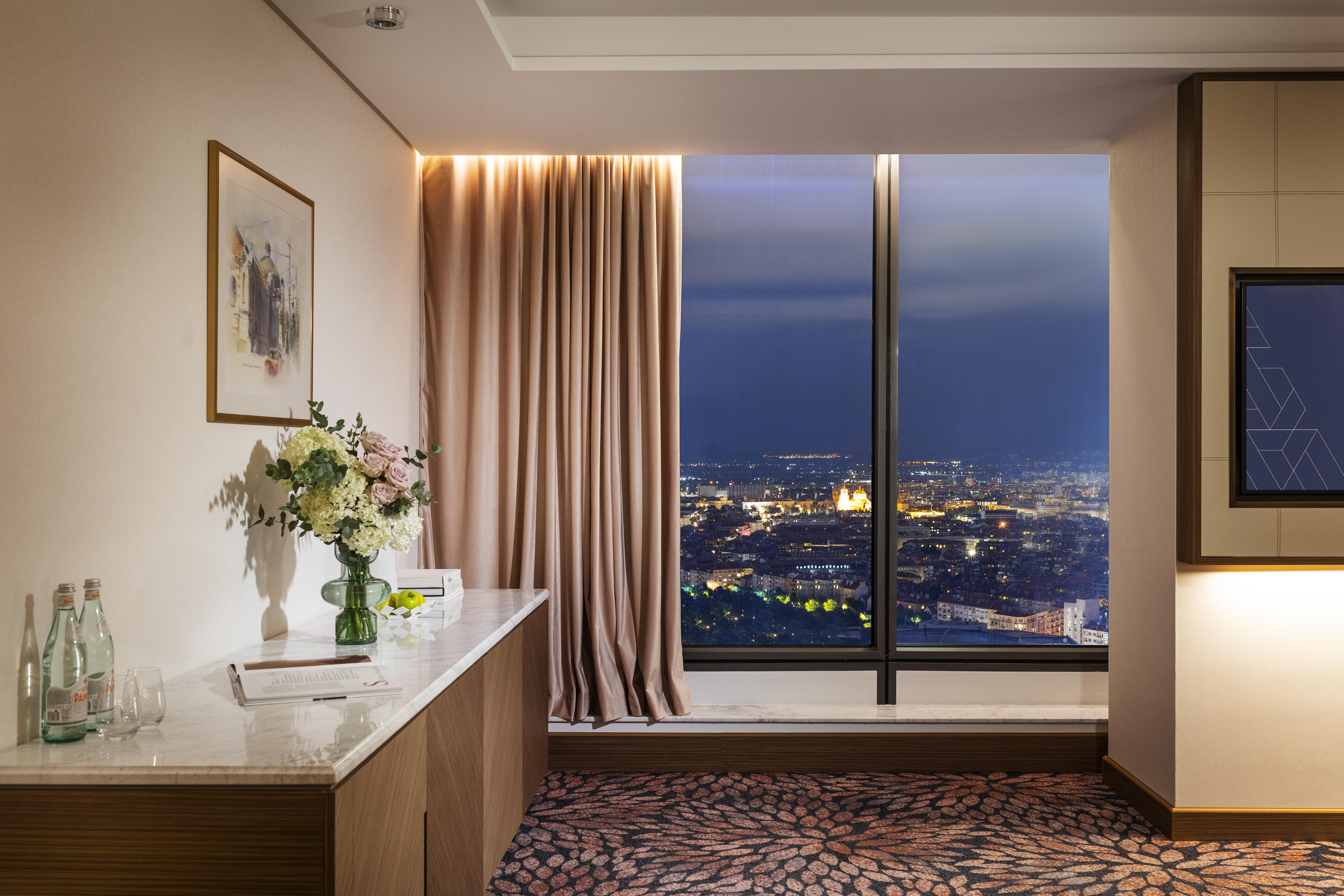 Grand Hotel Millennium 소피아 외부 사진 The photo depicts a modern hotel room or a lounge area with a stunning view of a cityscape at night. On the left, there's a wooden console table with a vase of flowers and a bottle of water. A piece of art is framed on the wall above the table. The r