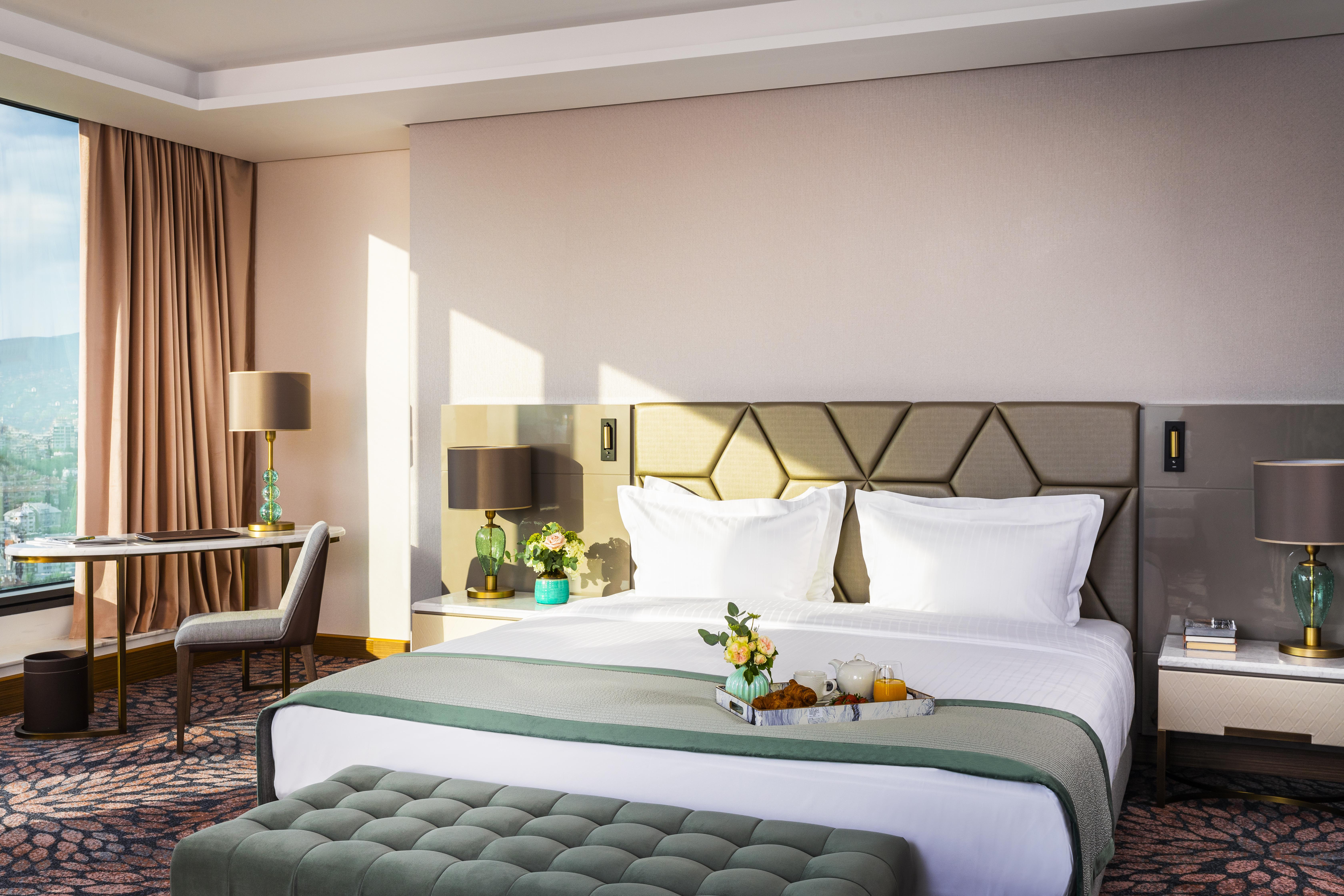 Grand Hotel Millennium 소피아 외부 사진 The photo depicts a stylish hotel room featuring a large bed with white linens and a green decorative throw. Behind the bed is a geometric-patterned headboard. To the side, there are modern bedside lamps and a small table with a vase of flowers. A se