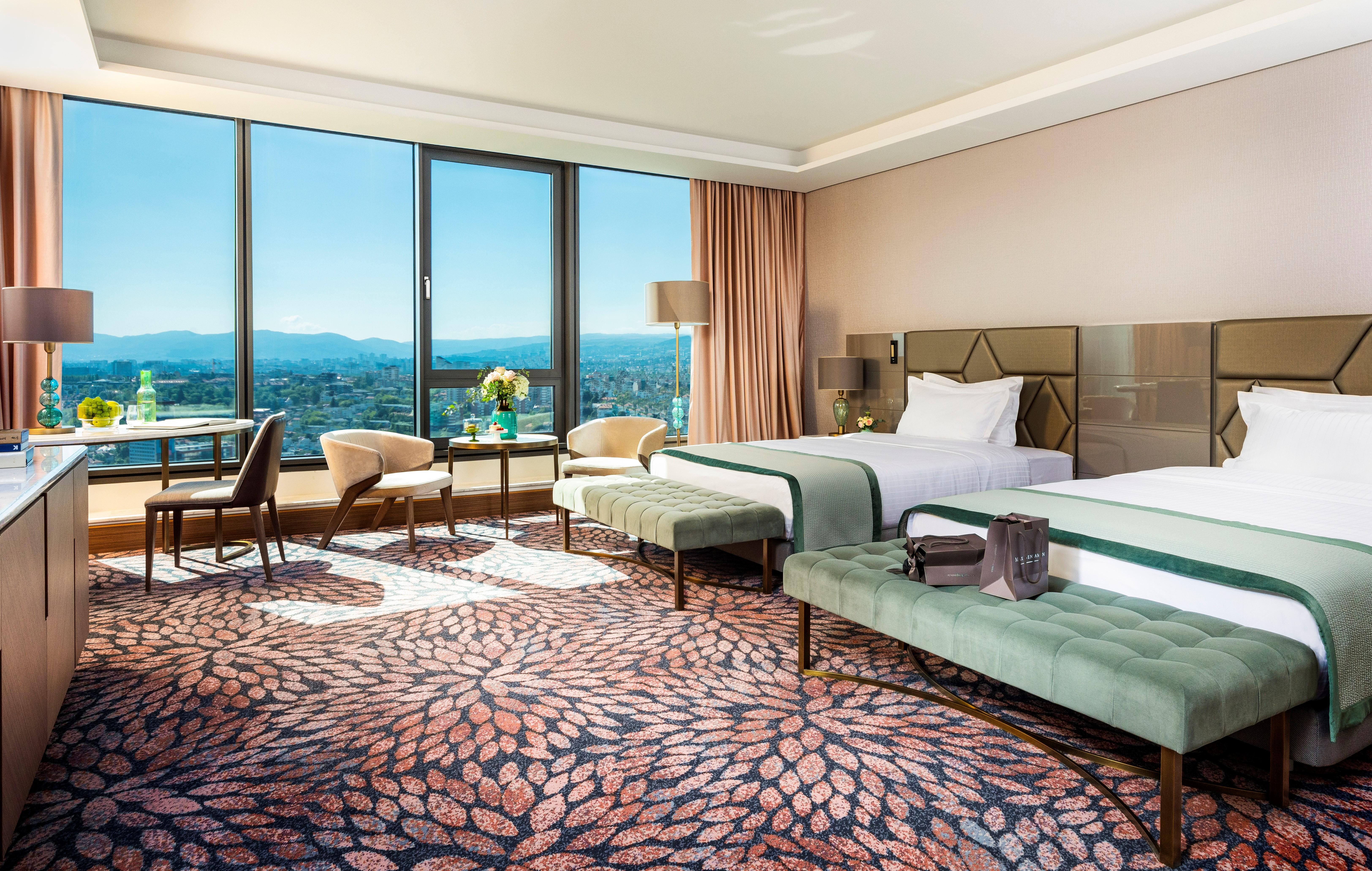 Grand Hotel Millennium 소피아 외부 사진 The photo depicts a luxurious hotel room featuring two beds with elegant bedding. The room has large windows that offer a panoramic view of the city and mountains in the background. There is a decorative area rug with a floral pattern covering the fl