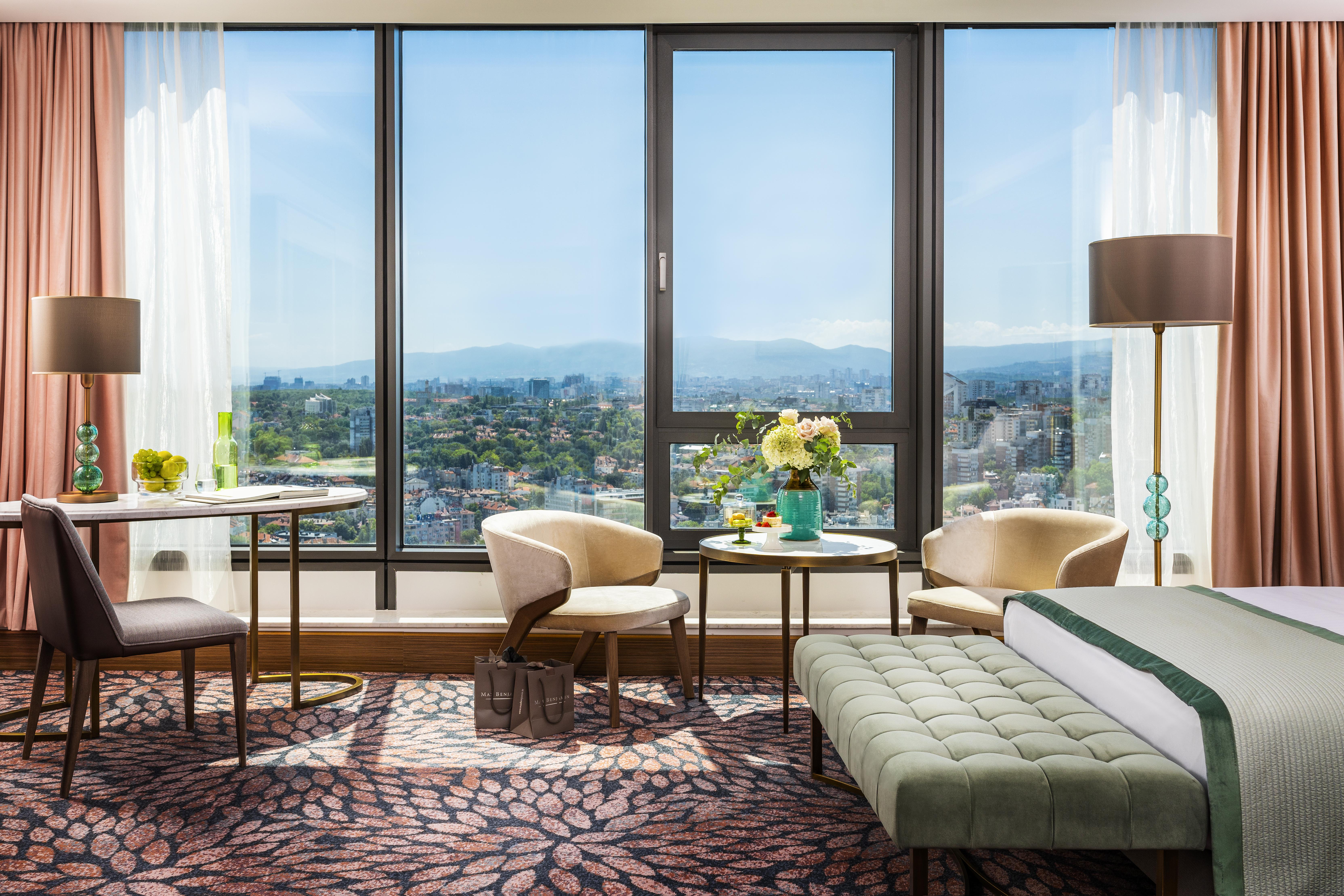 Grand Hotel Millennium 소피아 외부 사진 The photo shows a modern and well-designed hotel room featuring a large window that offers a view of the outdoors, including mountains in the background. Inside the room, there is a neatly made bed with a green accent blanket, a stylish rug with a fl