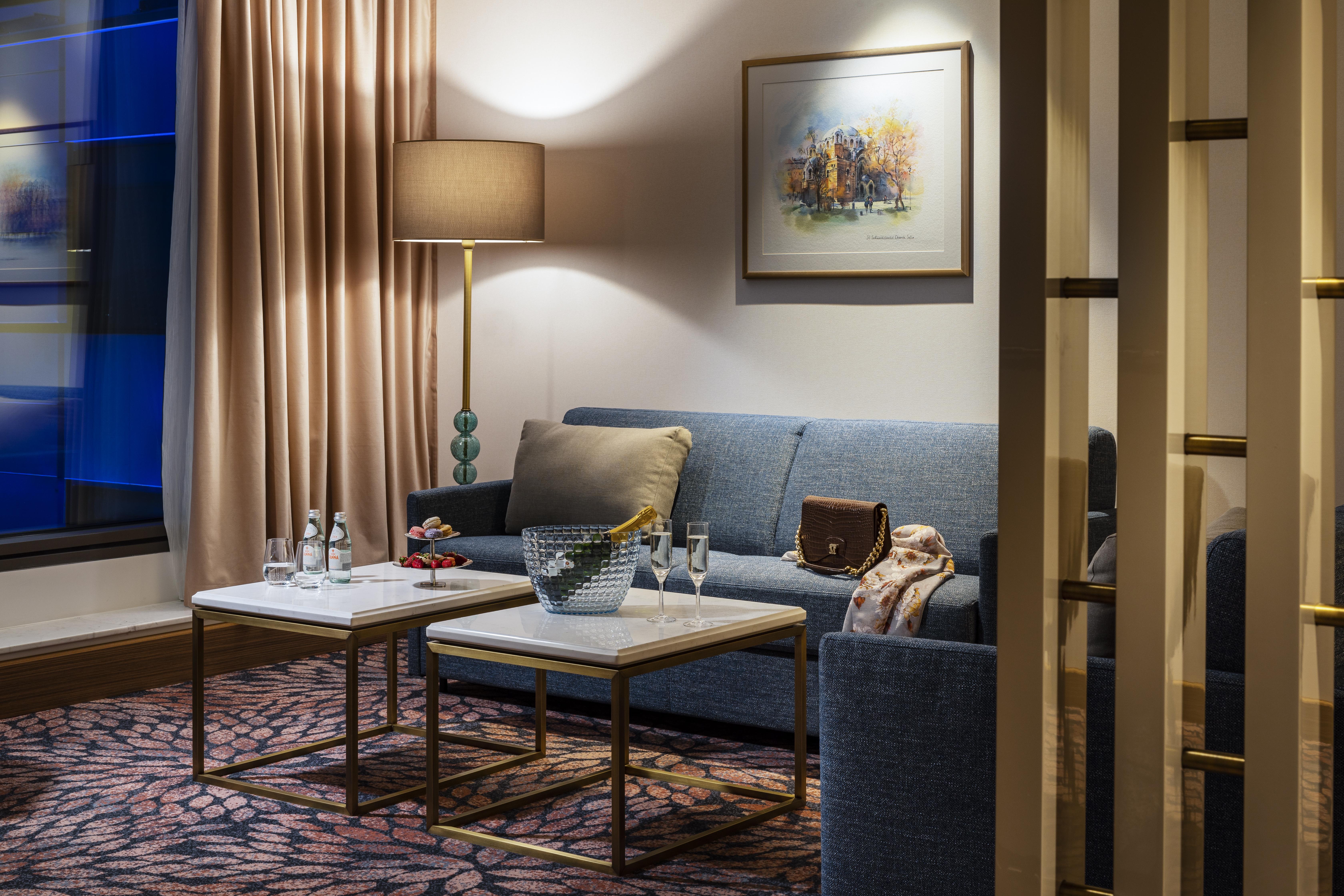Grand Hotel Millennium 소피아 외부 사진 The photo shows a cozy and elegantly decorated living space. It features a blue sofa against a wall, complemented by a painting above it. There are two small marble-top coffee tables with gold frames, one of which holds decorative items and glasses. 