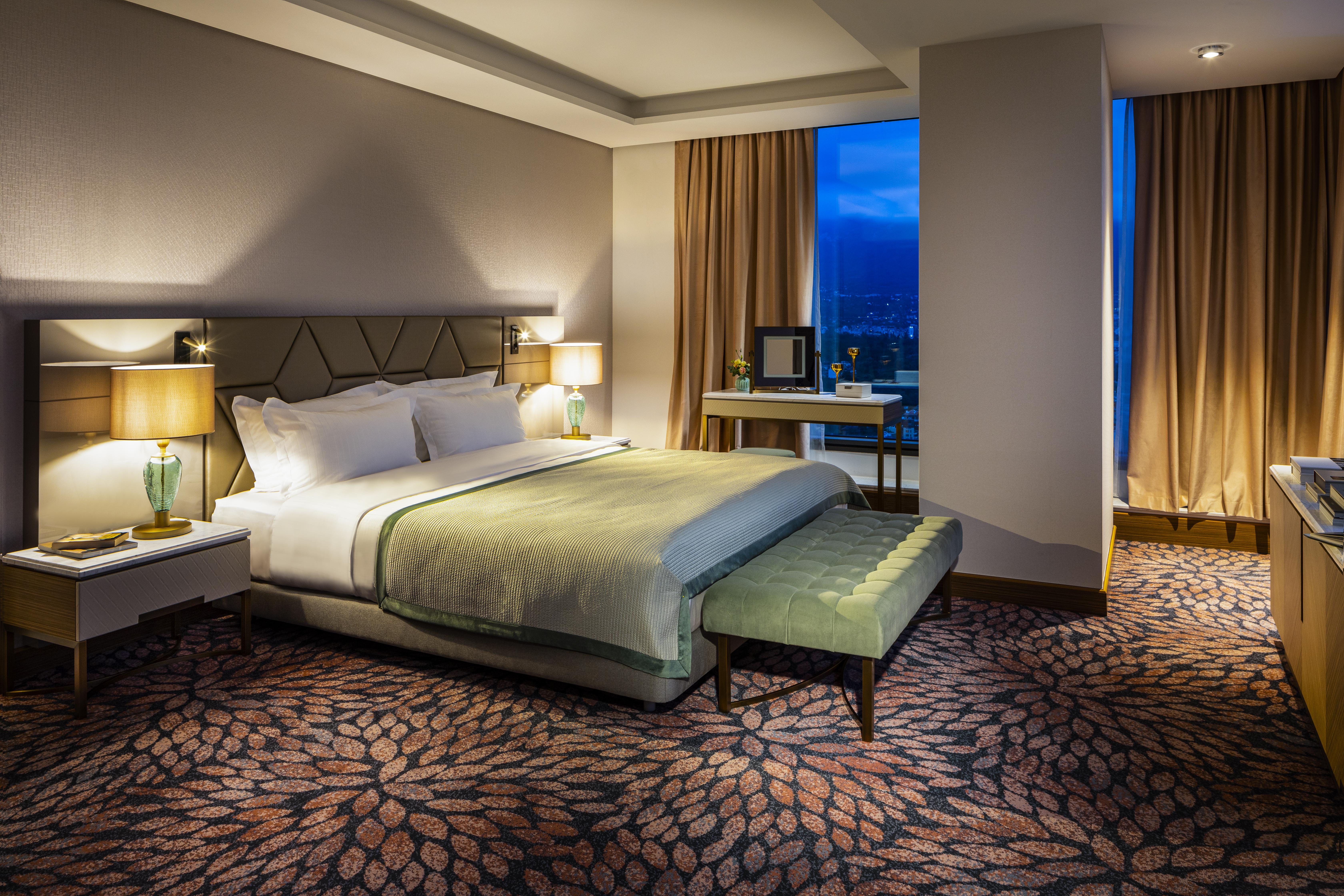 Grand Hotel Millennium 소피아 외부 사진 The photo shows a modern hotel room featuring a spacious design. It has a large bed with a light green throw blanket positioned in the center. On either side of the bed, there are stylish bedside tables with lamps. 

The floor has a patterned carpet,