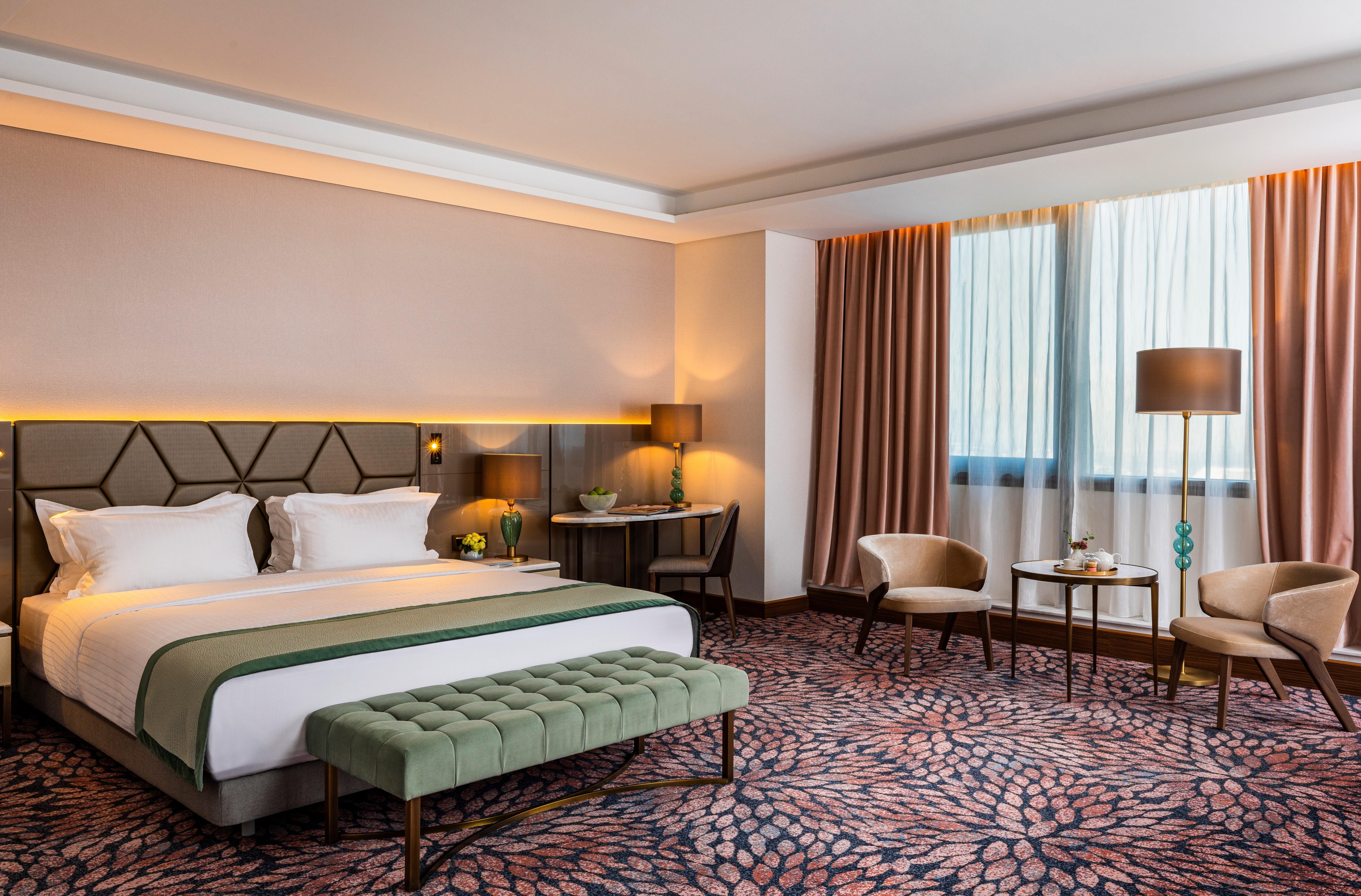 Grand Hotel Millennium 소피아 외부 사진 The photo shows a modern hotel room. It features a large bed with a headboard, topped with two white pillows and a green throw blanket. In front of the bed, there is a stylish green bench. On either side of the bed, there are bedside tables with lamp