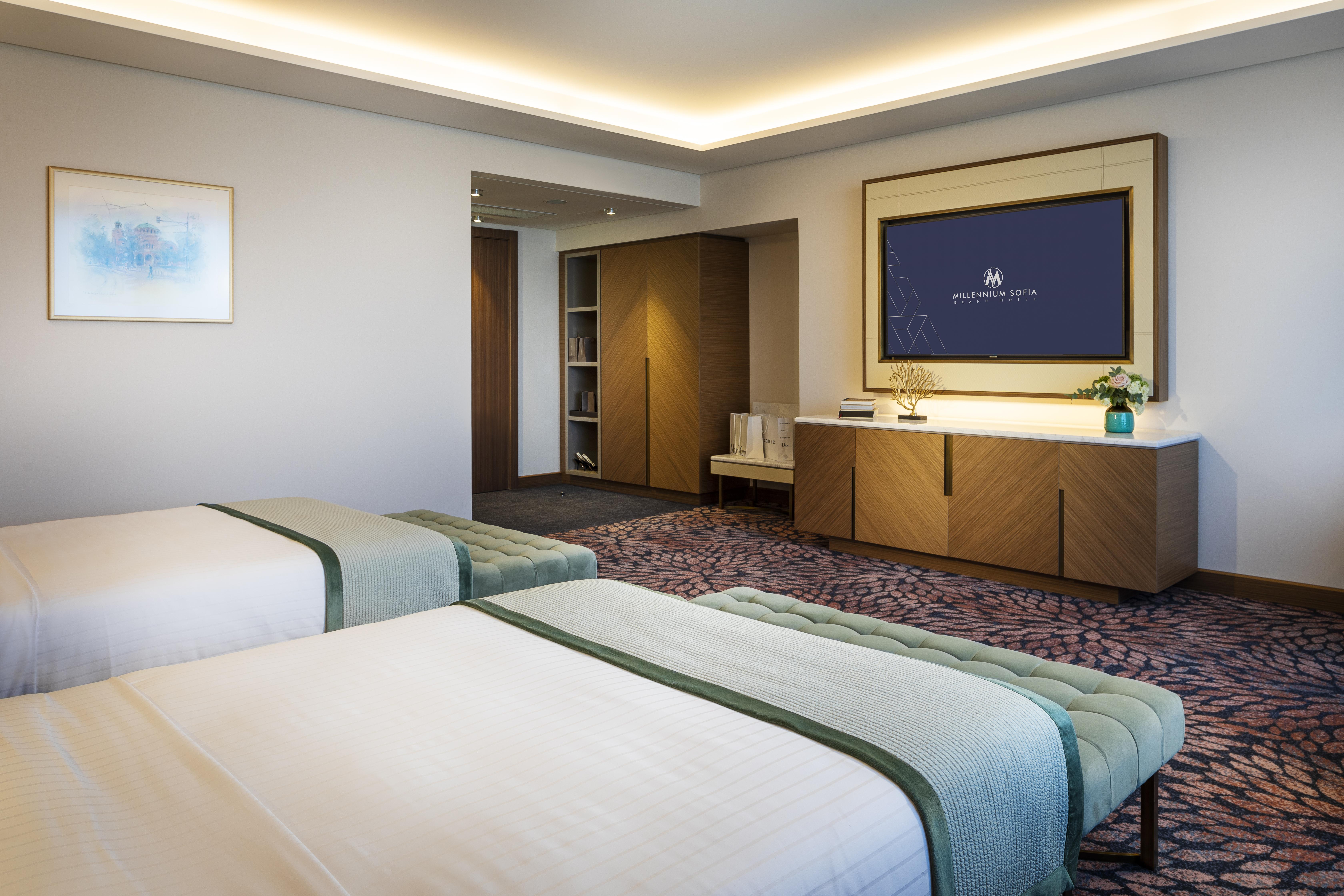 Grand Hotel Millennium 소피아 외부 사진 The photo shows a modern hotel room featuring two beds with light-colored bedding and decorative throws. The room has a contemporary design, characterized by neutral walls and a plush carpet. A large television is mounted on the wall across from the 