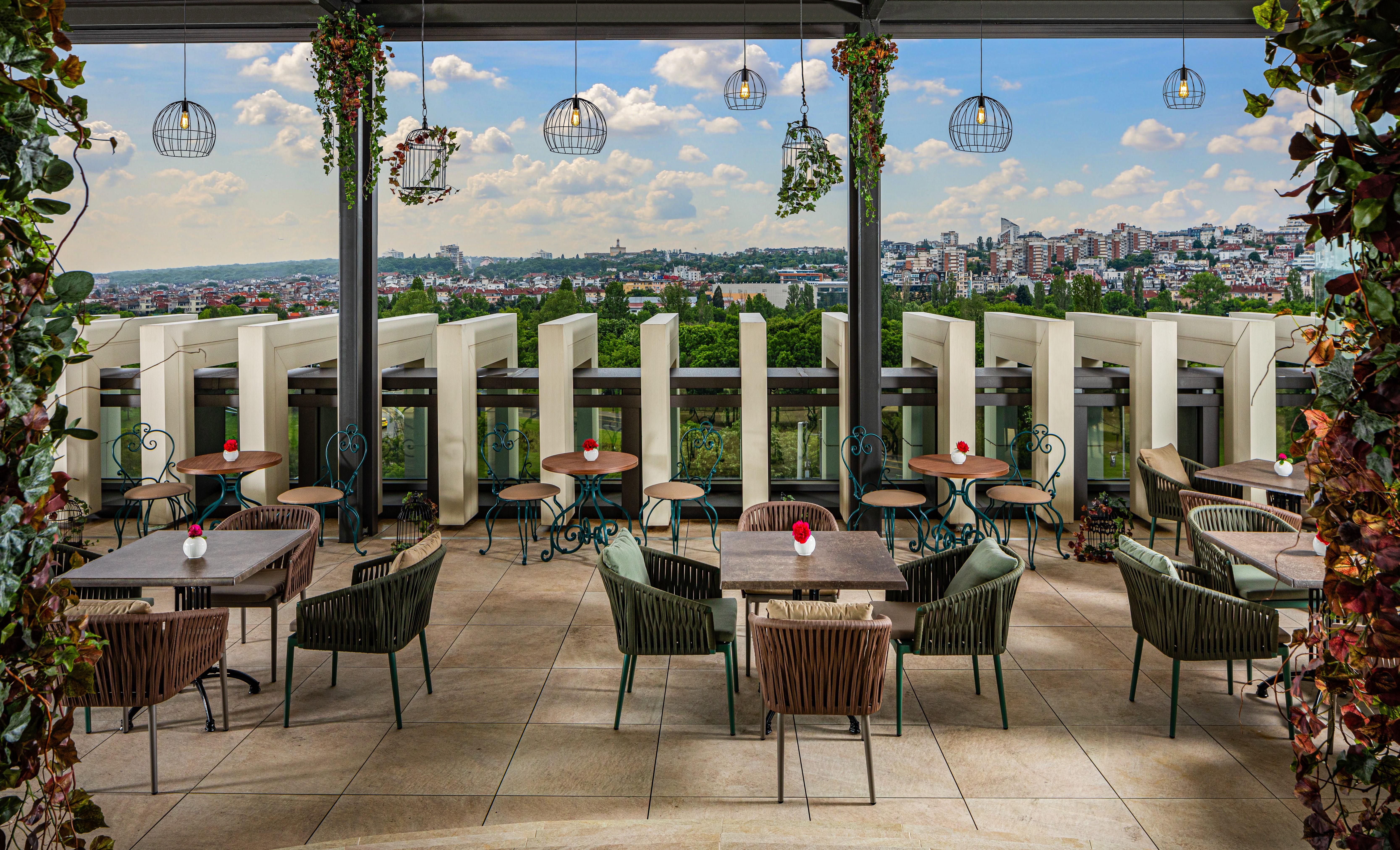 Grand Hotel Millennium 소피아 외부 사진 The photo shows a stylish outdoor restaurant or café setting. There are several tables arranged on a tiled patio, each with chairs, some made of woven materials. Centerpieces with small red flowers adorn each table. The space is partially shaded by o