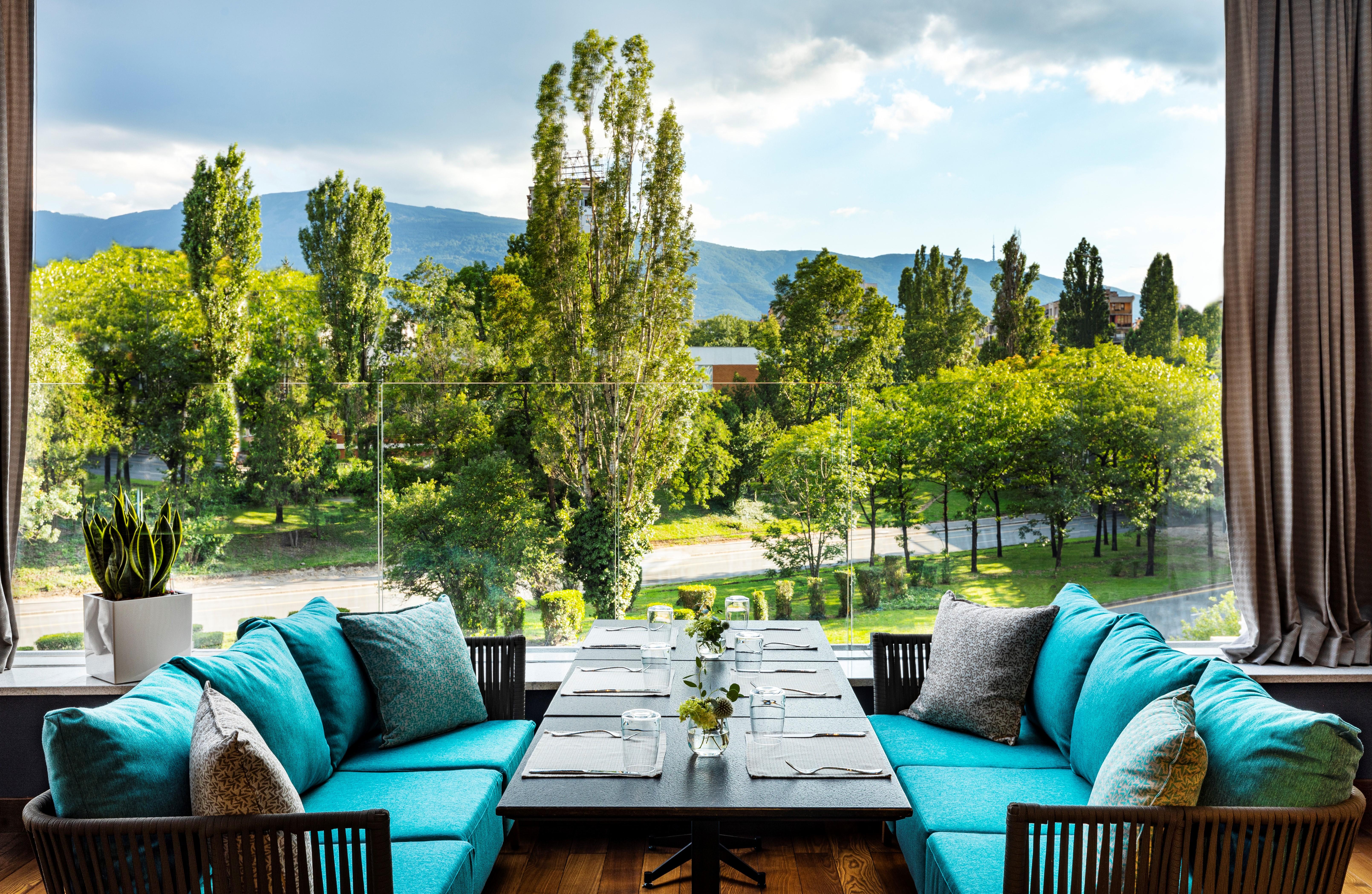 Grand Hotel Millennium 소피아 외부 사진 The photo shows a bright and inviting dining area with large windows that overlook a lush green landscape. There are several trees in the background, along with a view of distant mountains under a partly cloudy sky. The seating consists of stylish tu