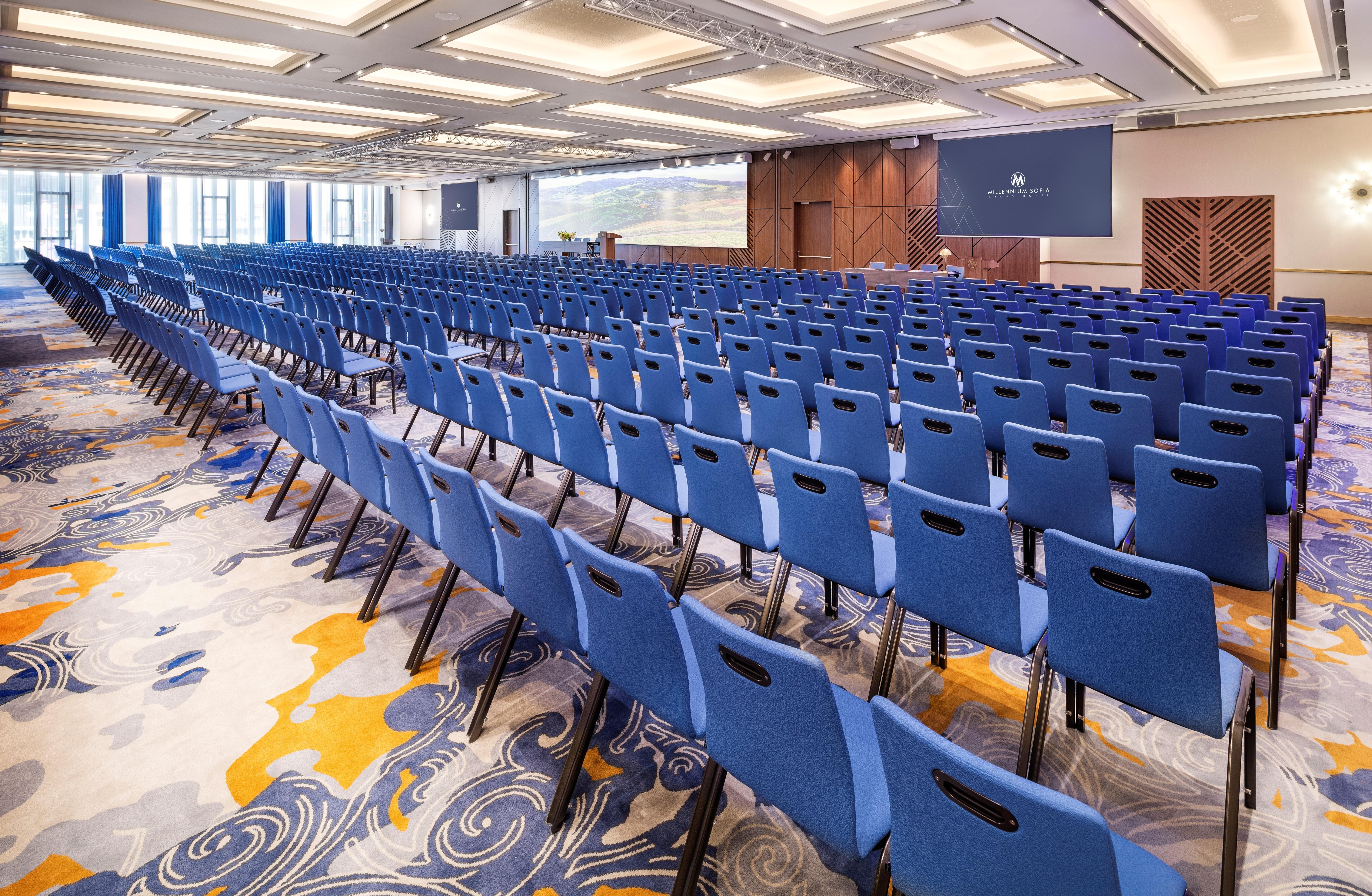 Grand Hotel Millennium 소피아 외부 사진 The photo shows a large conference or meeting room set up for an event. There are numerous blue chairs arranged in rows, creating a seating area for attendees. The floor features a decorative pattern, and the room has a modern design with ample light