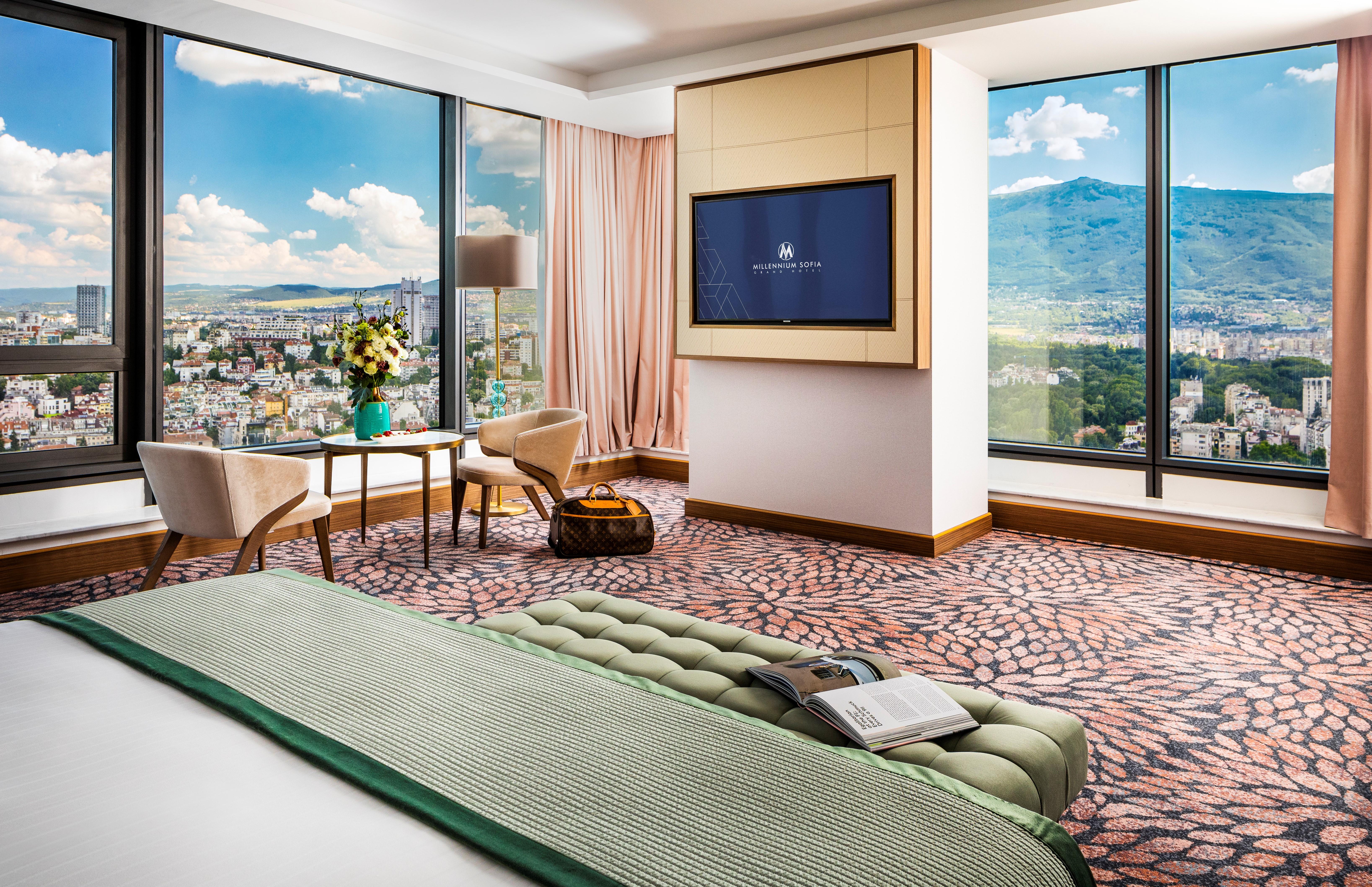 Grand Hotel Millennium 소피아 외부 사진 The photo showcases a stylish hotel room with modern decor. It features large windows providing a panoramic view of a cityscape and mountains in the background. Inside, there is a comfortable bed with a green bedspread, a seating area with two armcha
