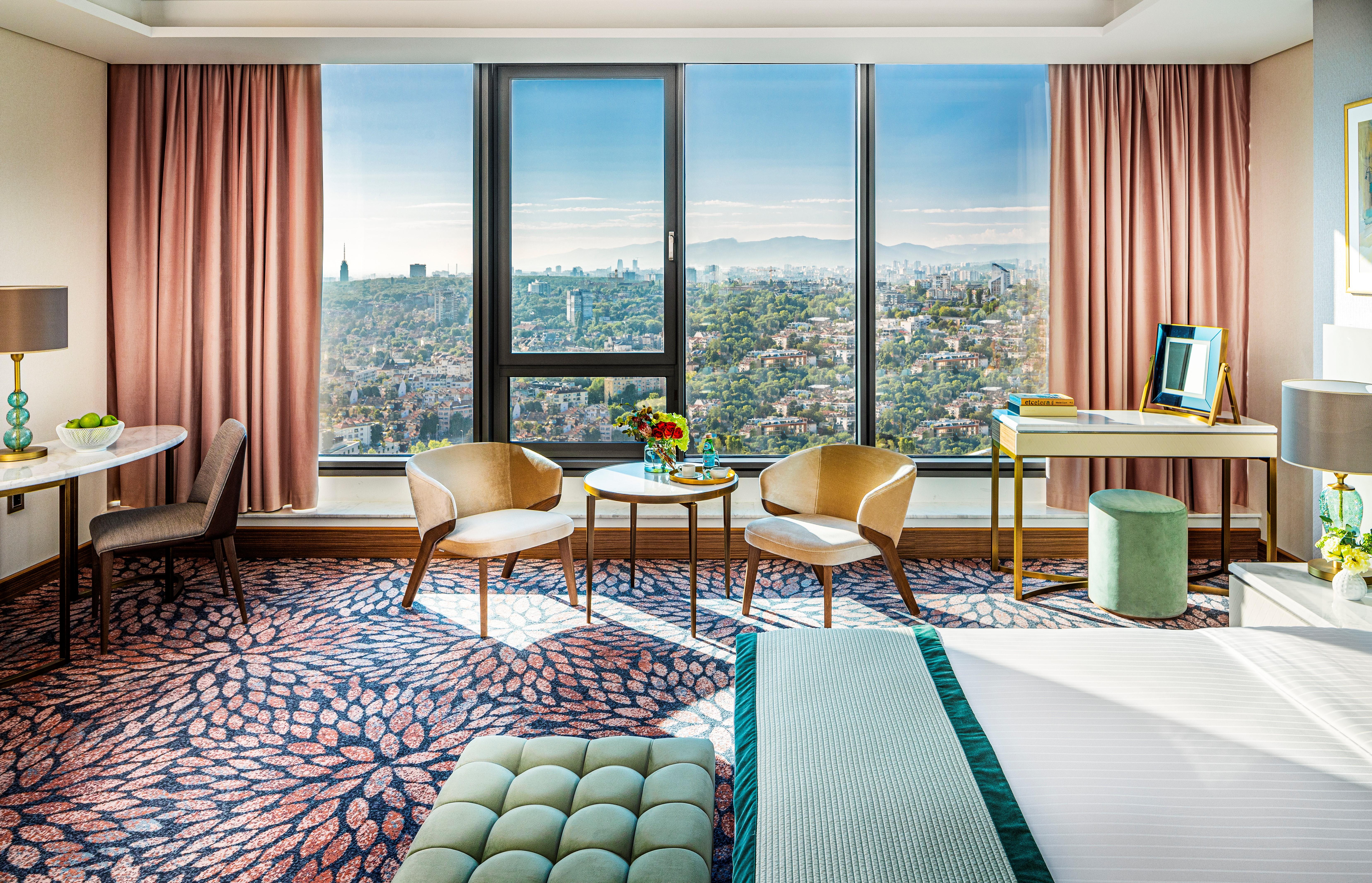 Grand Hotel Millennium 소피아 외부 사진 The photo shows a stylish hotel room featuring modern decor. There are large windows that offer a panoramic view of the cityscape, allowing natural light to fill the space. The walls are adorned with soft pink curtains. In the room, there are two ele