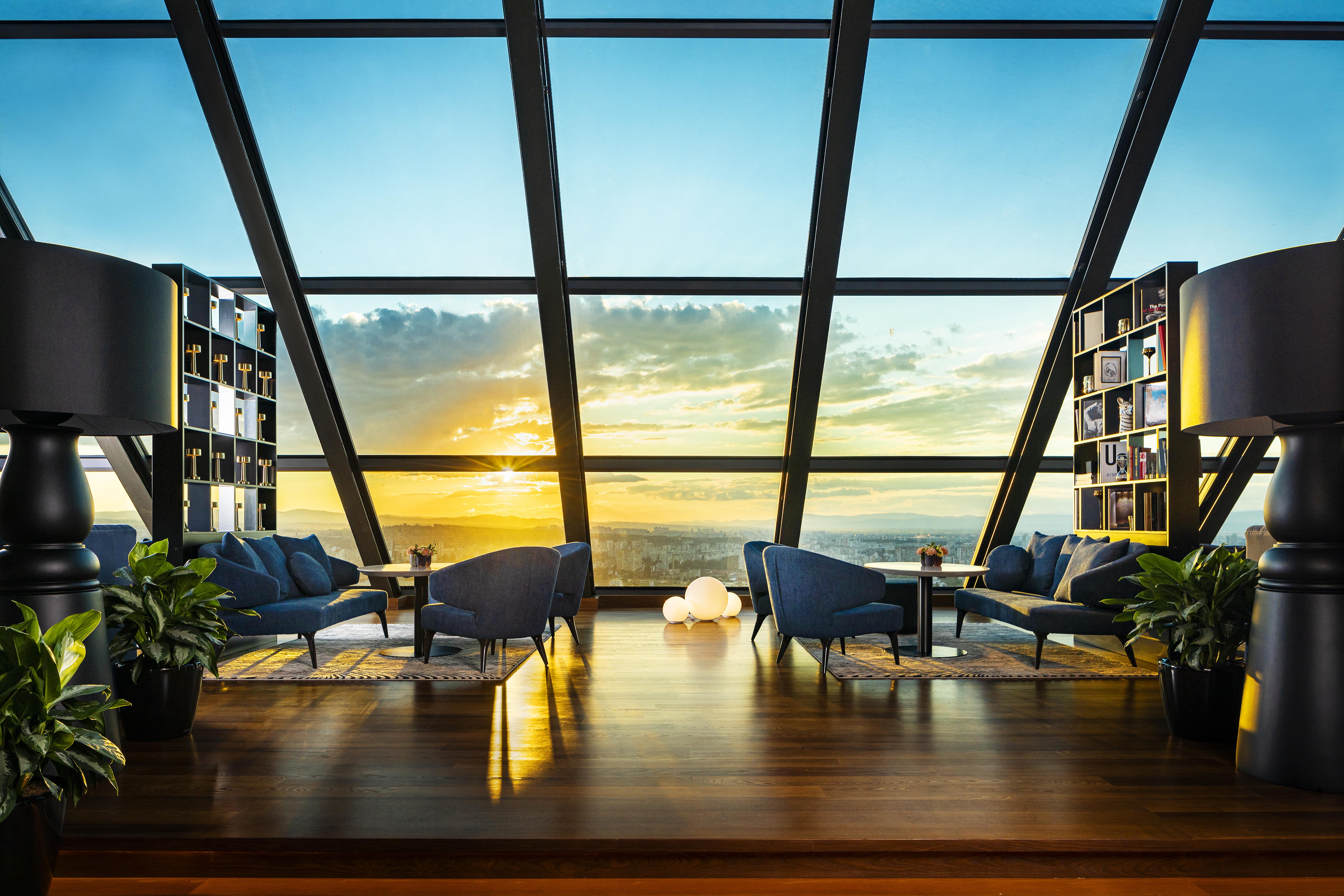 Grand Hotel Millennium 소피아 외부 사진 The photo depicts a modern, stylish interior of a high-rise space with a panoramic view through large glass windows. The scene features a cozy seating area with several blue armchairs arranged around a low table, creating a comfortable atmosphere. Th