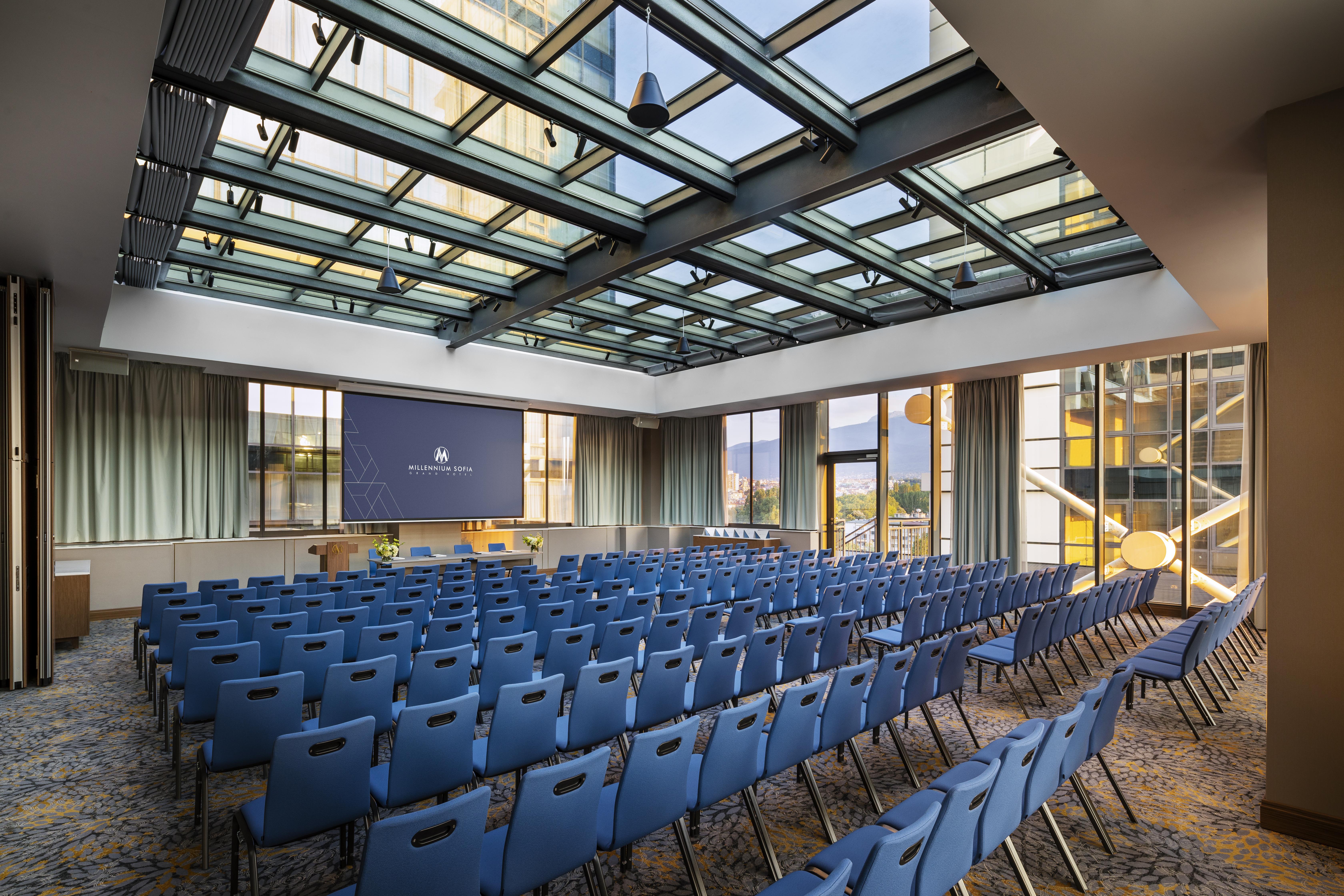 Grand Hotel Millennium 소피아 외부 사진 The photo shows a large conference room or meeting space. It features rows of blue chairs arranged in a theater style, facing a large screen at the front of the room. The room is well-lit, with a glass ceiling allowing natural light to filter in. The
