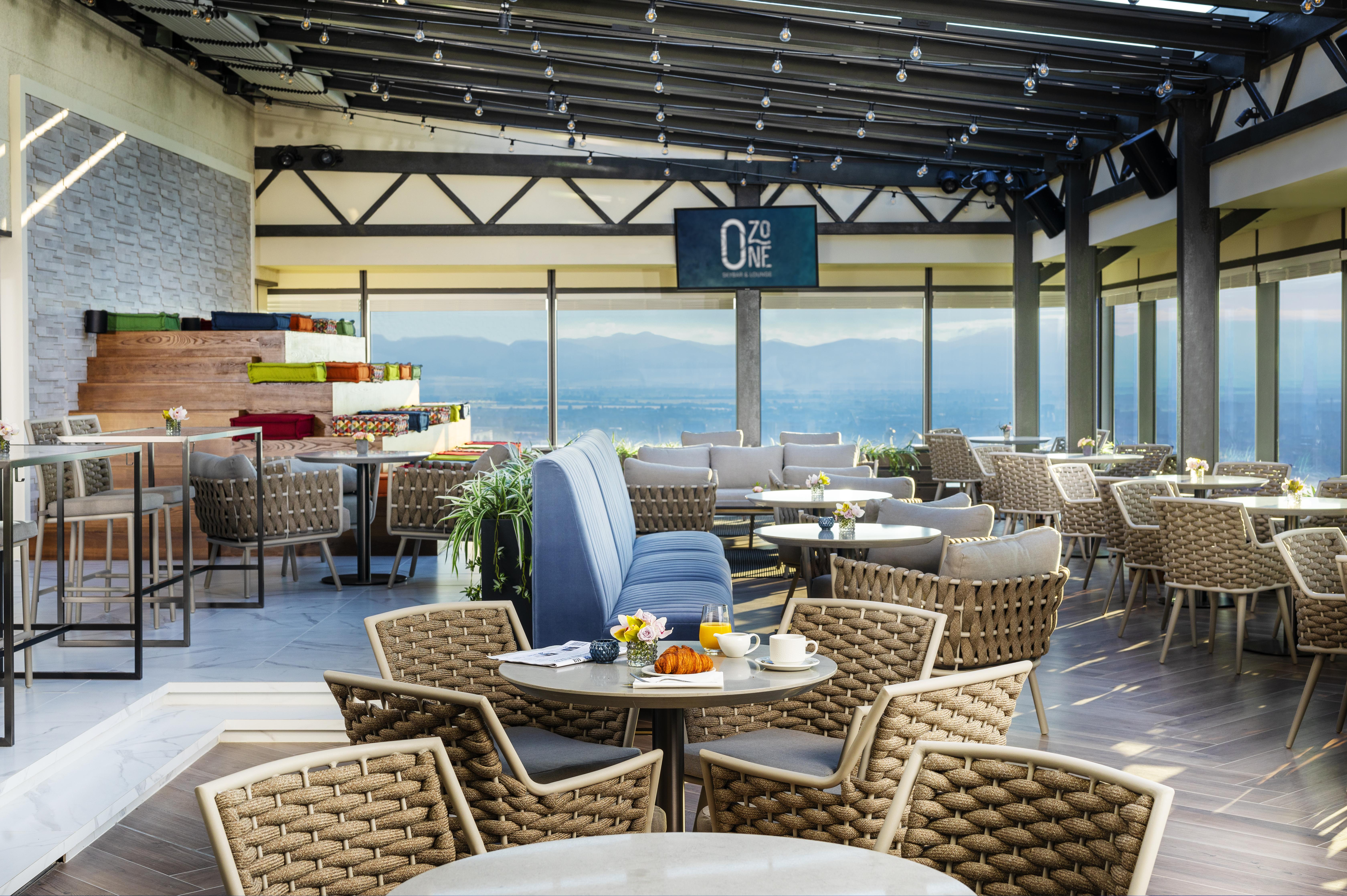Grand Hotel Millennium 소피아 외부 사진 The photo shows a modern restaurant or café with a spacious interior. It features a variety of seating arrangements, including round tables surrounded by wicker chairs and lounges. Large windows allow for a panoramic view, which likely includes sceni