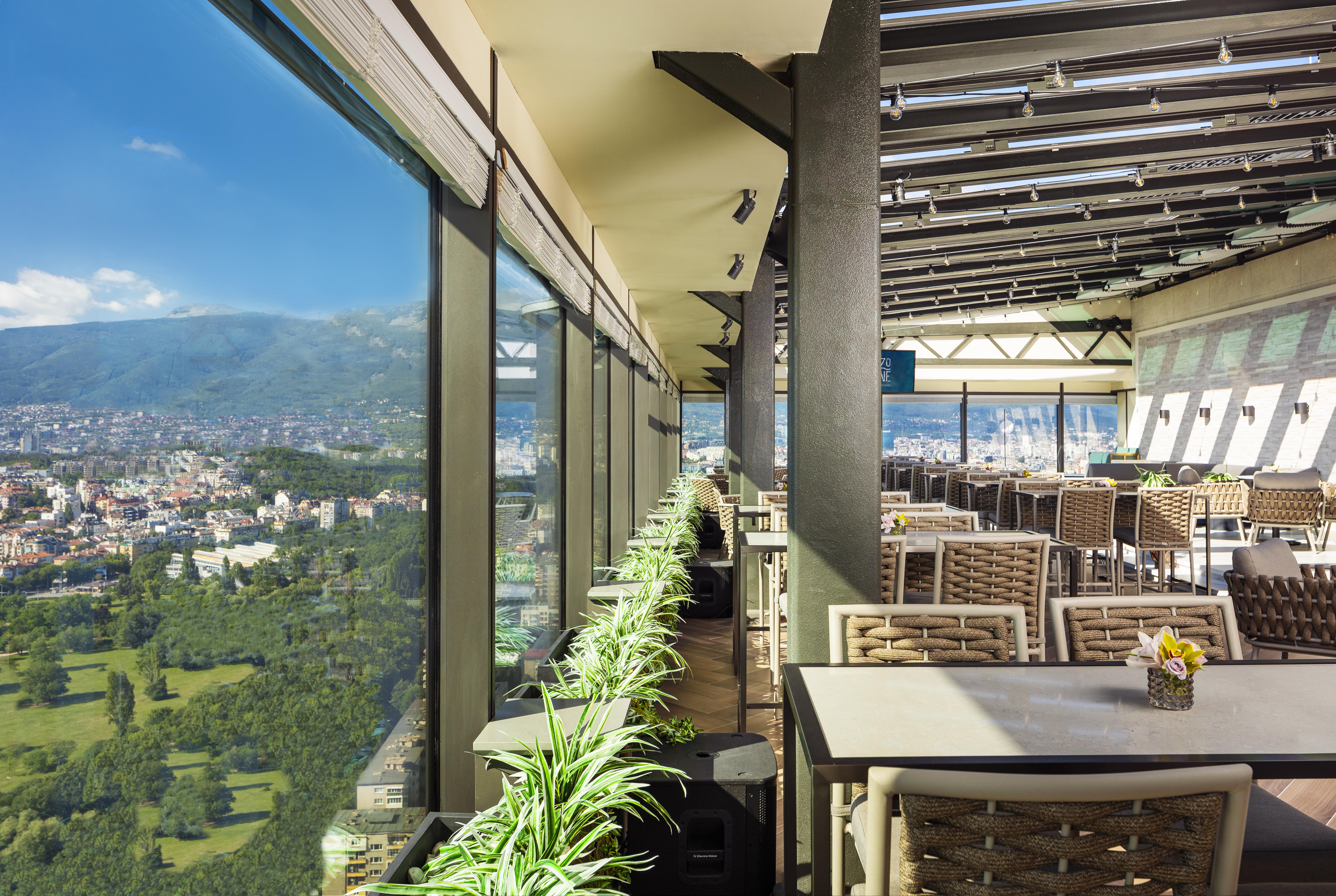 Grand Hotel Millennium 소피아 외부 사진 The photo shows a modern, spacious restaurant with large glass windows offering a panoramic view of a city and surrounding landscape. The interior features contemporary furniture, including stylish tables and chairs. There are potted plants along the